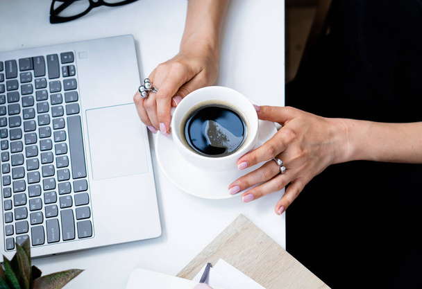 Жінки рука тримає чашку кави в затишному літньому офісі з ноутбуком і зеленими рослинами
 - Фото, зображення