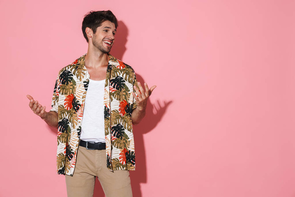 Portrait of cheerful young man in flamboyant shirt smiling and posing on camera isolated over pink background - Photo, image