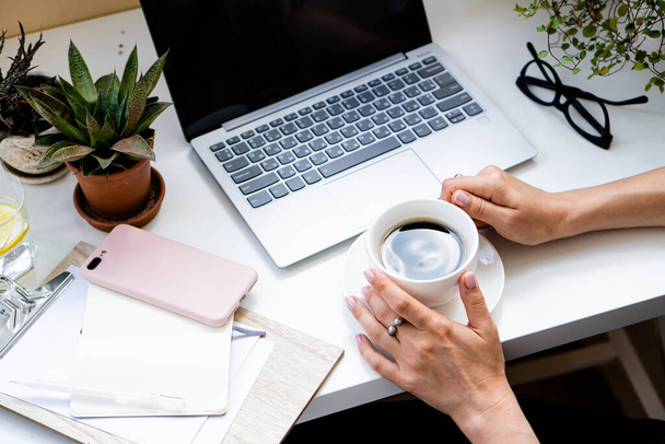 Mãos femininas segurando xícara de café no escritório acolhedor com laptop e plantas verdes
 - Foto, Imagem