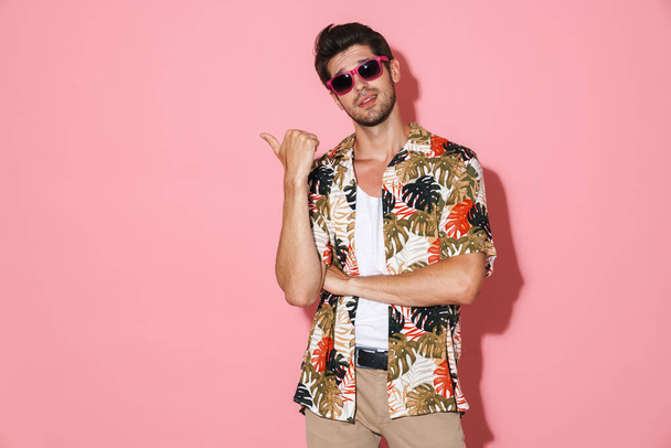 Portrait of handsome young man in sunglasses pointing fingers aside isolated over pink background - Photo, image