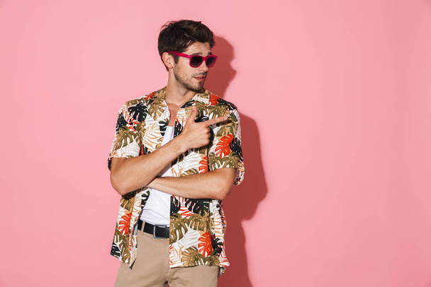 Portrait of handsome young man in sunglasses pointing fingers aside isolated over pink background - Fotoğraf, Görsel