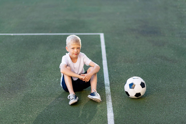 um menino loiro de uniforme esportivo se senta em um campo de futebol com uma bola de futebol, seção de esportes. Treinamento de crianças, lazer de crianças
. - Foto, Imagem