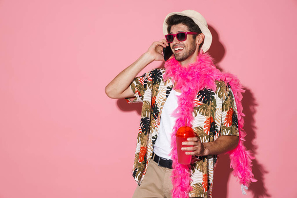 Portrait of smiling man wearing boa talking on cellphone while drinking soda isolated over pink background - Фото, изображение