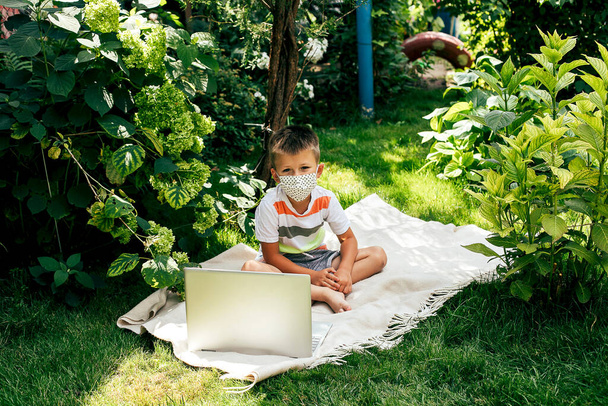 Maskeli küçük bir çocuk, karantina döneminde dizüstü bilgisayarla çimlerin üzerinde yatıyordu.. - Fotoğraf, Görsel
