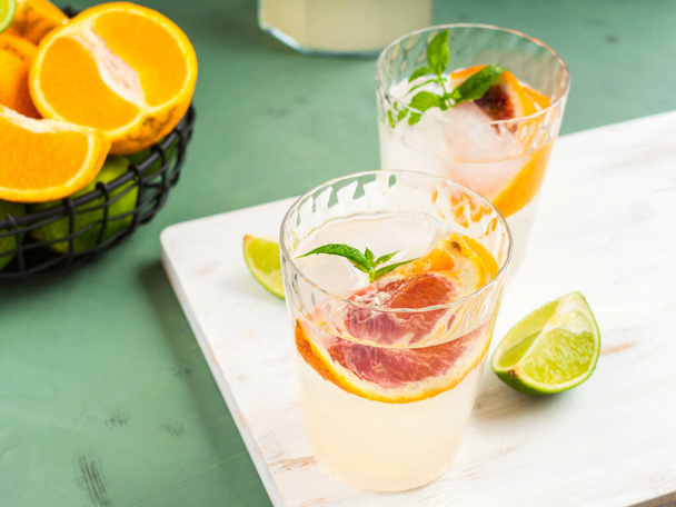 Limonata di agrumi fresca estiva con lime, menta, arance rosse e cubetti di ghiaccio in vaso e bicchieri su sfondo verde strutturato. - Foto, immagini