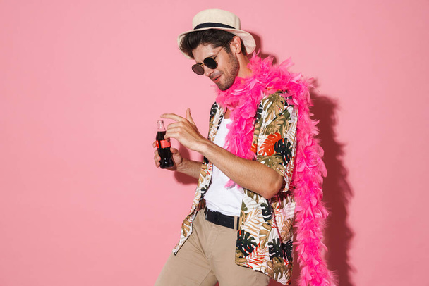 Portrait of cheerful young man wearing boa dancing while drinking soda isolated over pink background - Valokuva, kuva