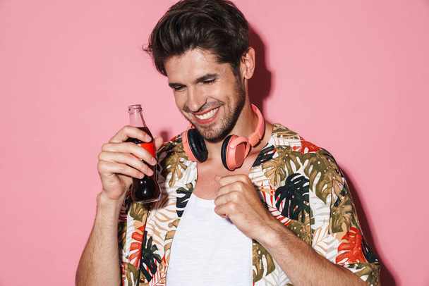 Retrato de un joven alegre usando auriculares inalámbricos mientras bebe soda aislada sobre fondo rosa - Foto, Imagen