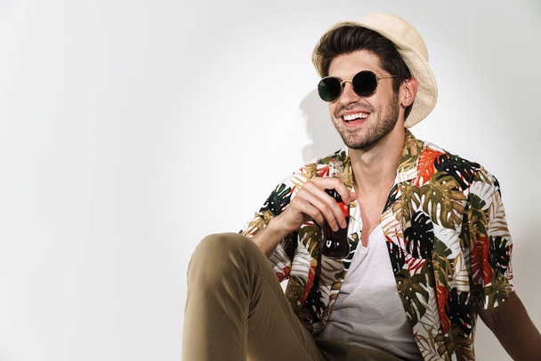 Porträt eines fröhlichen jungen Mannes mit Sonnenbrille, der Limo trinkt, während er isoliert auf dem Boden vor weißem Hintergrund sitzt - Foto, Bild
