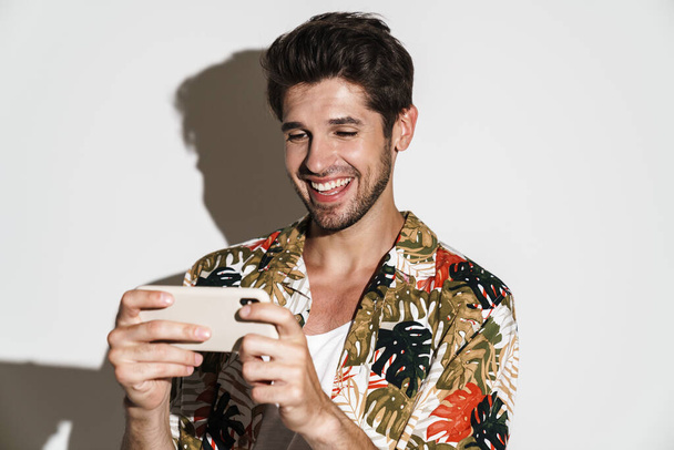 Portrait of cheerful young man in flamboyant shirt playing online game on cellphone isolated over white background - Photo, image