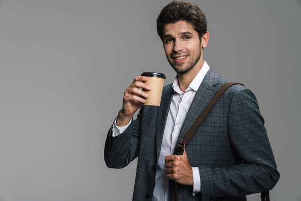 Foto di un uomo d'affari sorridente e sicuro di sé con borsa che beve caffè da asporto isolato sul muro grigio - Foto, immagini