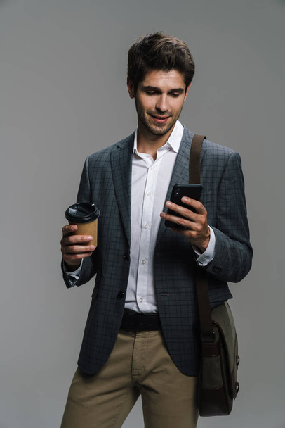 Photo of smiling confident businessman drinking coffee takeaway and using cellphone isolated over grey wall - Foto, Imagem