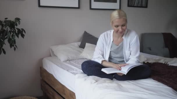 piękna młoda blondynka dziewczyna czyta książkę siedzi na przytulnym łóżku. - Materiał filmowy, wideo