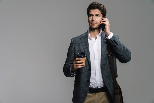 Photo of young confident businessman drinking coffee takeaway and talking on cellphone isolated over grey wall - Foto, afbeelding
