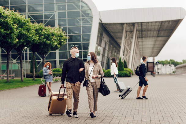 Jonge man en vrouw in beschermende maskers met een koffer gaan naar het vliegveld. Veilig reizen na een coronaviruspandemie. - Foto, afbeelding