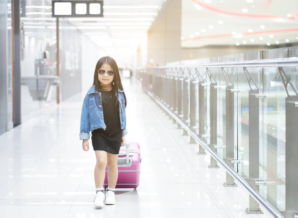 Азиатский ребенок или девочка ходить перетаскивая багаж или багаж и розовый чемодан с носить солнцезащитные очки и джинсы рубашку или джинсы на пешеходной дорожке в аэропорту для отдыха отдых и летние каникулы поездки на теплой - Фото, изображение