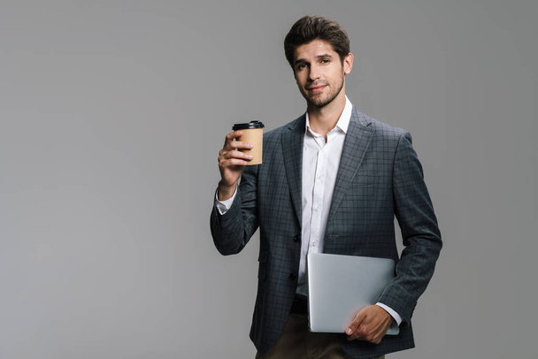 Photo of pleased unshaven businessman drinking coffee takeaway and holding laptop isolated over grey wall - Valokuva, kuva