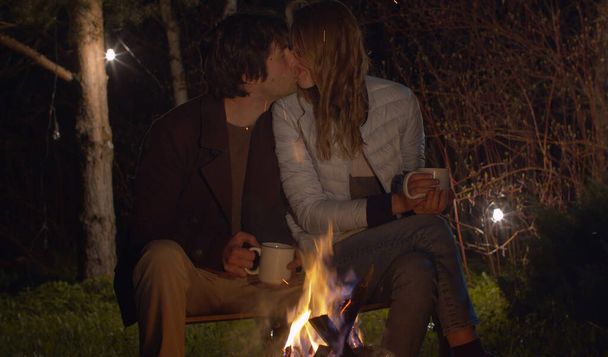 Young happy couple at night sitting near campfire and kissing. Family on weekend in the woods - 写真・画像