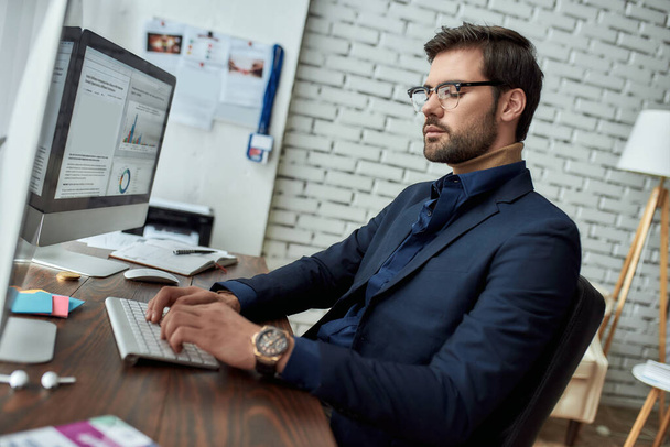 Visão lateral de um analista financeiro usando óculos trabalhando com dados estatísticos enquanto estava sentado em seu local de trabalho no escritório moderno
 - Foto, Imagem