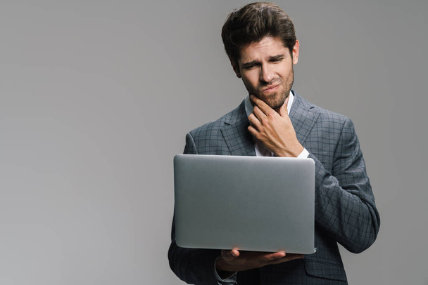 Photo of displeased young businessman working with laptop isolated over grey wall - Foto, Imagem