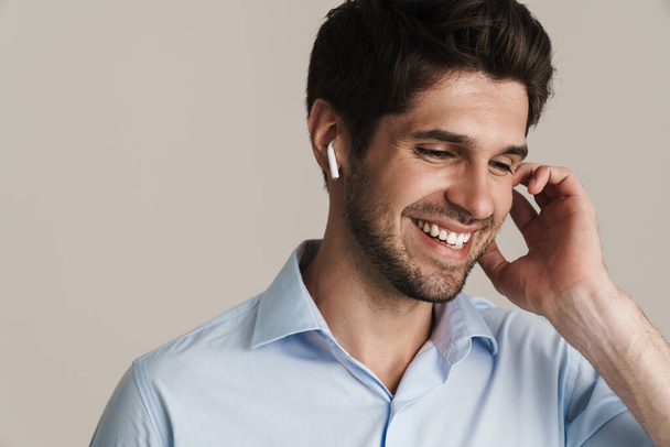 Portrait of cheerful handsome man laughing while using wireless earphone isolated over white background - Photo, Image