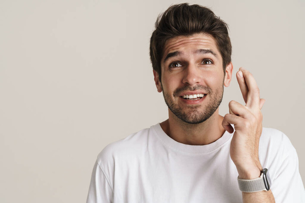 Portrait of pleased young man smiling while holding fingers crossed for good luck isolated over white background - Photo, image