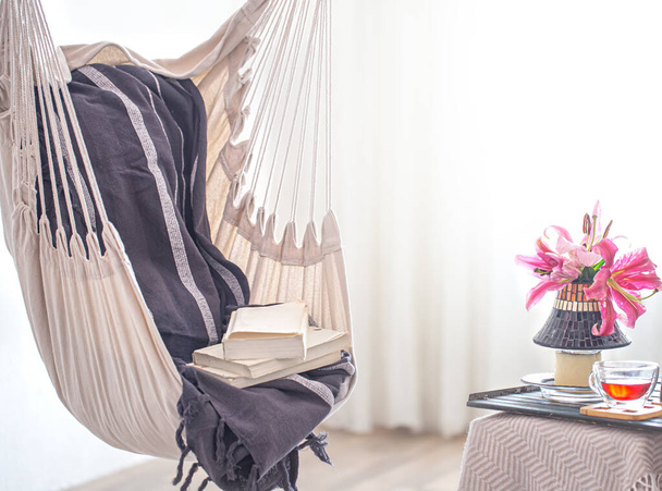 Een boho-stijl hangmat stoel met een stapel boeken. Houten dienblad met theepot en kopje thee en mooie Lily bloemen. Gezellig interieur. Het concept van rust en comfort thuis. Ruimte voor tekst. - Foto, afbeelding