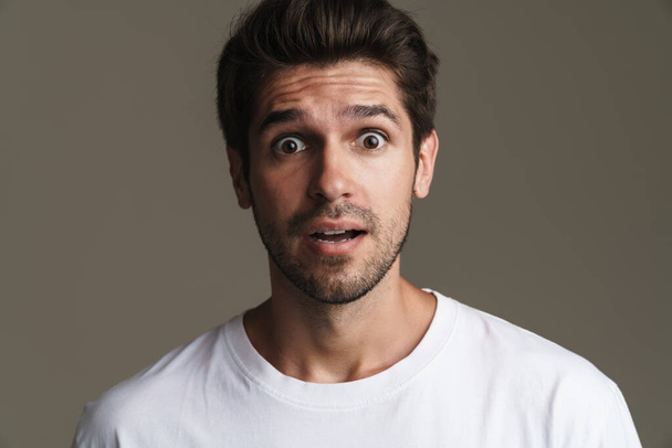 Portrait of shocked young man in basic t-shirt expressing surprise on camera isolated over grey background - Foto, imagen