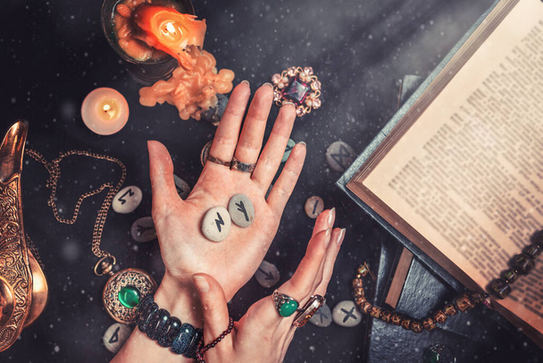 Astrologie und Esoterik. Die Hexe hält die Runensteine in den Händen. Auf schwarzem Hintergrund liegen wahrsagende Runen, ein Buch, kostbare Amulette, eine Kupferlampe und eine Kerze. Staub und Licht, - Foto, Bild