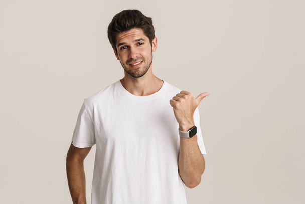 Portrait of pleased young man in basic t-shirt smiling and pointing finger aside isolated over white background - Fotografie, Obrázek