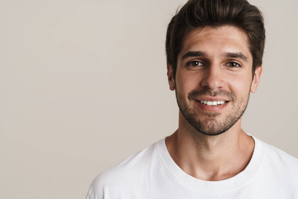 Portrait of joyful young man in basic t-shirt smiling and looking at camera isolated over white background - Zdjęcie, obraz