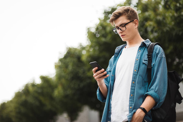 Νεαρός σύγχρονος φοιτητής συνομιλίας στο δίκτυο, ενώ στέκεται στη μέση του δρόμου. Όμορφος τύπος με γυαλιά και ένα τηλέφωνο στα χέρια του. - Φωτογραφία, εικόνα