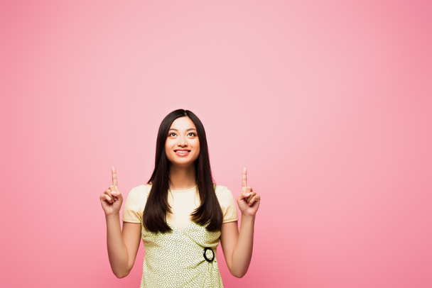 happy asian girl pointing with fingers and looking up isolated on pink  - Fotó, kép