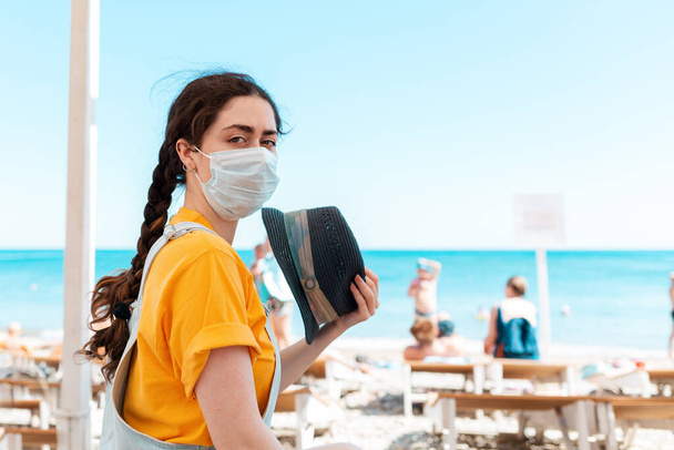 Een vrouw met een medisch masker, die zichzelf aanwakkert met een hoed van de hitte. Op de achtergrond, het strand en de zee. Het concept vakantie tijdens een virale pandemie. - Foto, afbeelding