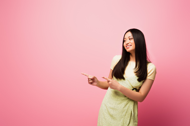 cheerful asian girl pointing with fingers and looking away on pink  - Foto, Bild