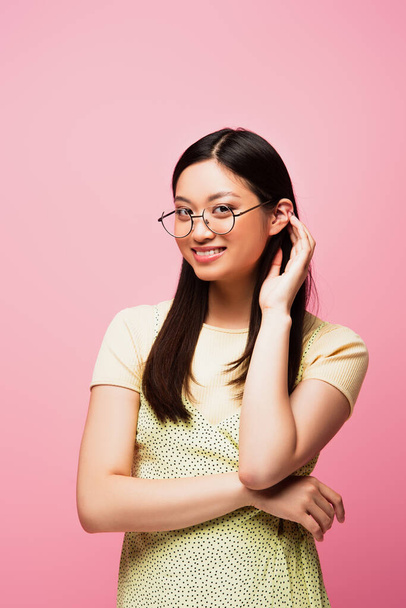 cheerful asian girl in glasses touching hair isolated on pink  - Фото, изображение