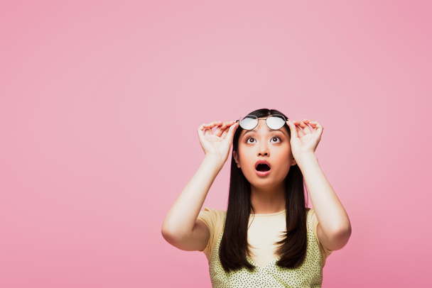 shocked asian girl looking up and touching glasses isolated on pink  - Zdjęcie, obraz