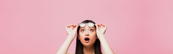 πανοραμική λήψη σοκαρισμένη Ασιάτισσα κοπέλα κοιτάζοντας ψηλά και αγγίζοντας τα γυαλιά που απομονώνονται σε ροζ  - Φωτογραφία, εικόνα