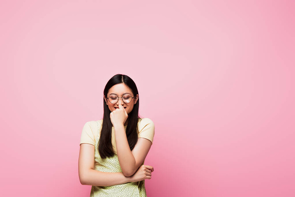 cheerful and young asian woman with closed eyes touching glasses isolated on pink - 写真・画像