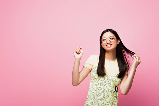 alegre e jovem asiático mulher no óculos tocando cabelo e olhando para longe isolado no rosa
 - Foto, Imagem