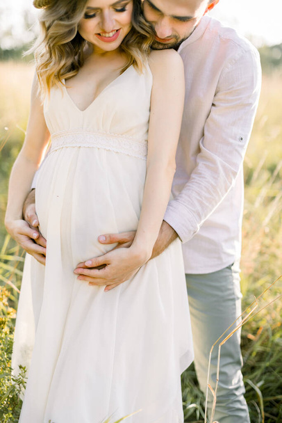 Cropped close up shot of happy man and his pregnant woman in white dress outdoors on a green summer field. Pregnancy and parenthood. Love and waiting for baby. Pregnant couple. - Photo, image