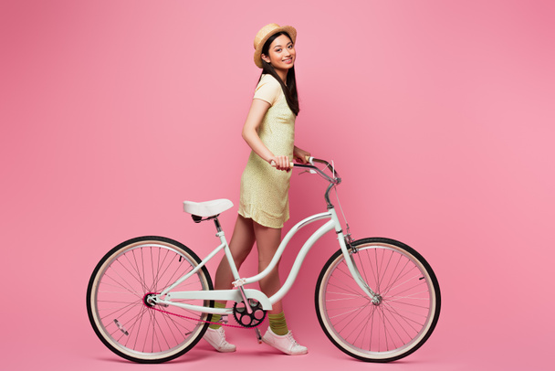 fröhliche asiatische junge Frau mit Strohhut geht mit Fahrrad auf rosa   - Foto, Bild