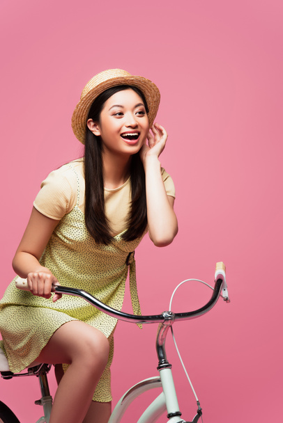 excited asian young woman in straw hat riding bicycle on pink   - Fotoğraf, Görsel