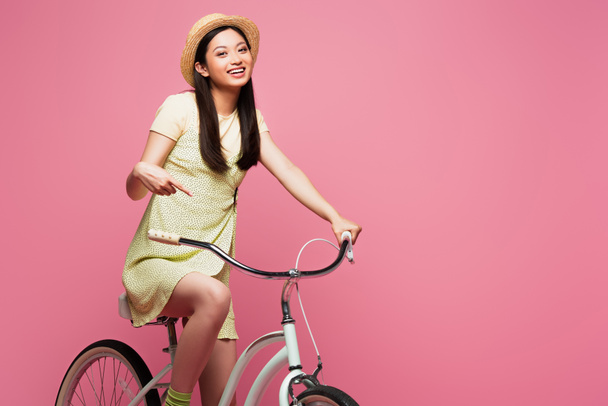 sorridente asiatico giovane donna in paglia cappello equitazione bicicletta isolato su rosa   - Foto, immagini