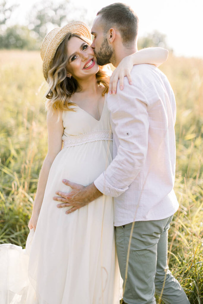 Waiting for the baby, pregnancy concept. Happy young couple, expecting a baby, walking in bright summer sunny day on a field. Man hugs and kisses his pretty pregnant woman in hat and dress - Photo, image