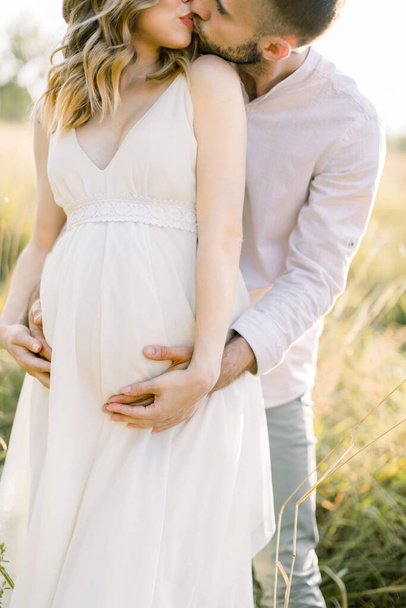 Cropped close up shot of happy man and his pregnant woman in white dress outdoors on a green summer field. Pregnancy and parenthood. Love and waiting for baby. Pregnant couple. - Foto, Bild