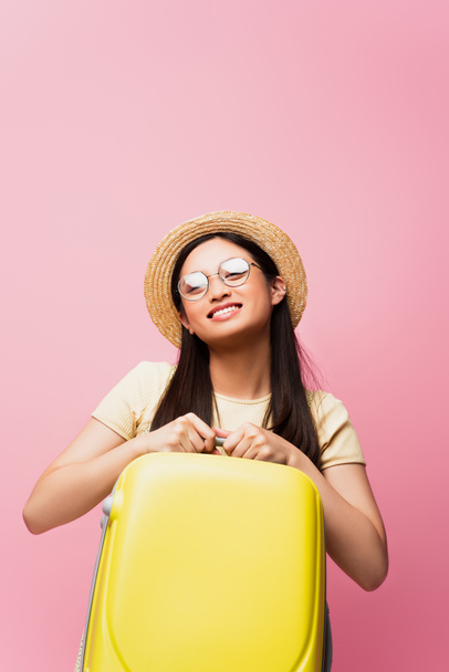 positive asian girl in glasses and straw hat holding yellow luggage on pink  - Valokuva, kuva