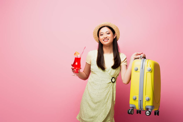 smiling asian woman in straw hat holding cocktail and yellow baggage on pink - Fotografie, Obrázek