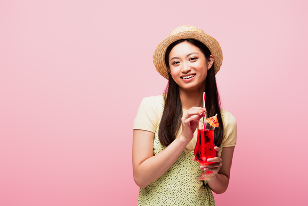 uśmiechnięta Azjatka w słomkowym kapeluszu trzymająca szklankę z koktajlem wyizolowanym na różowo - Zdjęcie, obraz