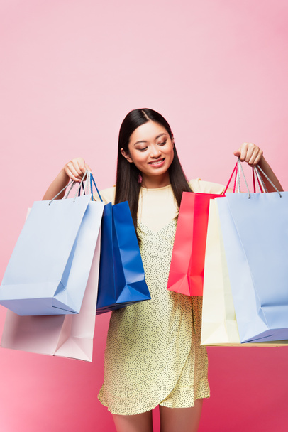 cheerful asian girl looking at shopping bags on pink  - Photo, image