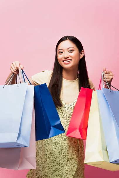 cheerful asian girl looking at camera and holding shopping bags isolated on pink  - Φωτογραφία, εικόνα